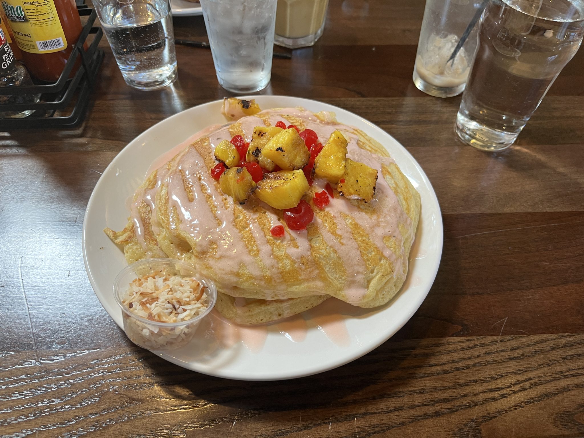 Pancakes with toasted pineapple, maraschino cherries and cherry sauce on top. A small cup of toasted coconut next to them.