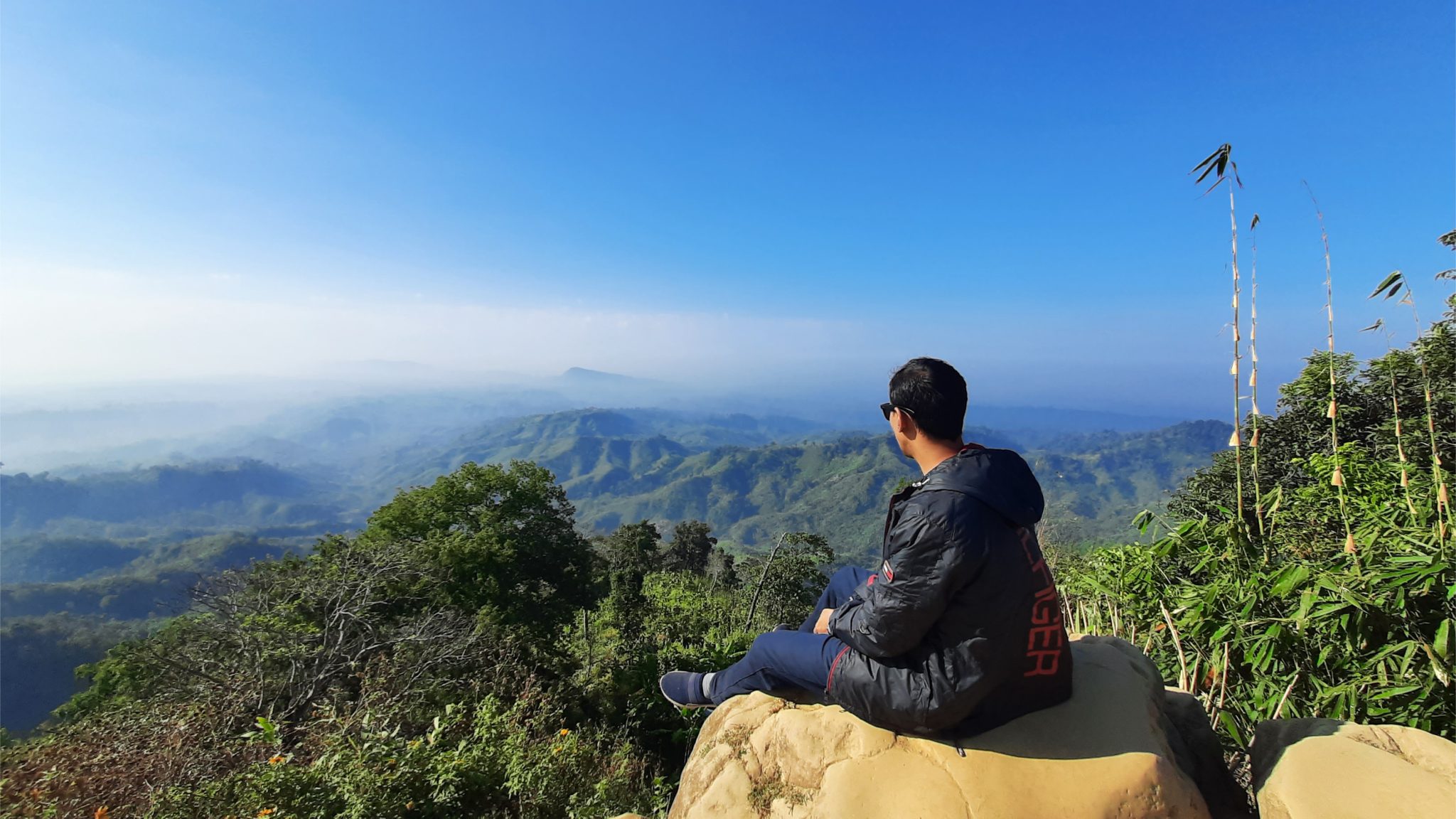 Person sitting on a rock on a mountain top looking over a vast expanse of forested mountains shrouded in clouds.  Sajek, Bangladesh.
