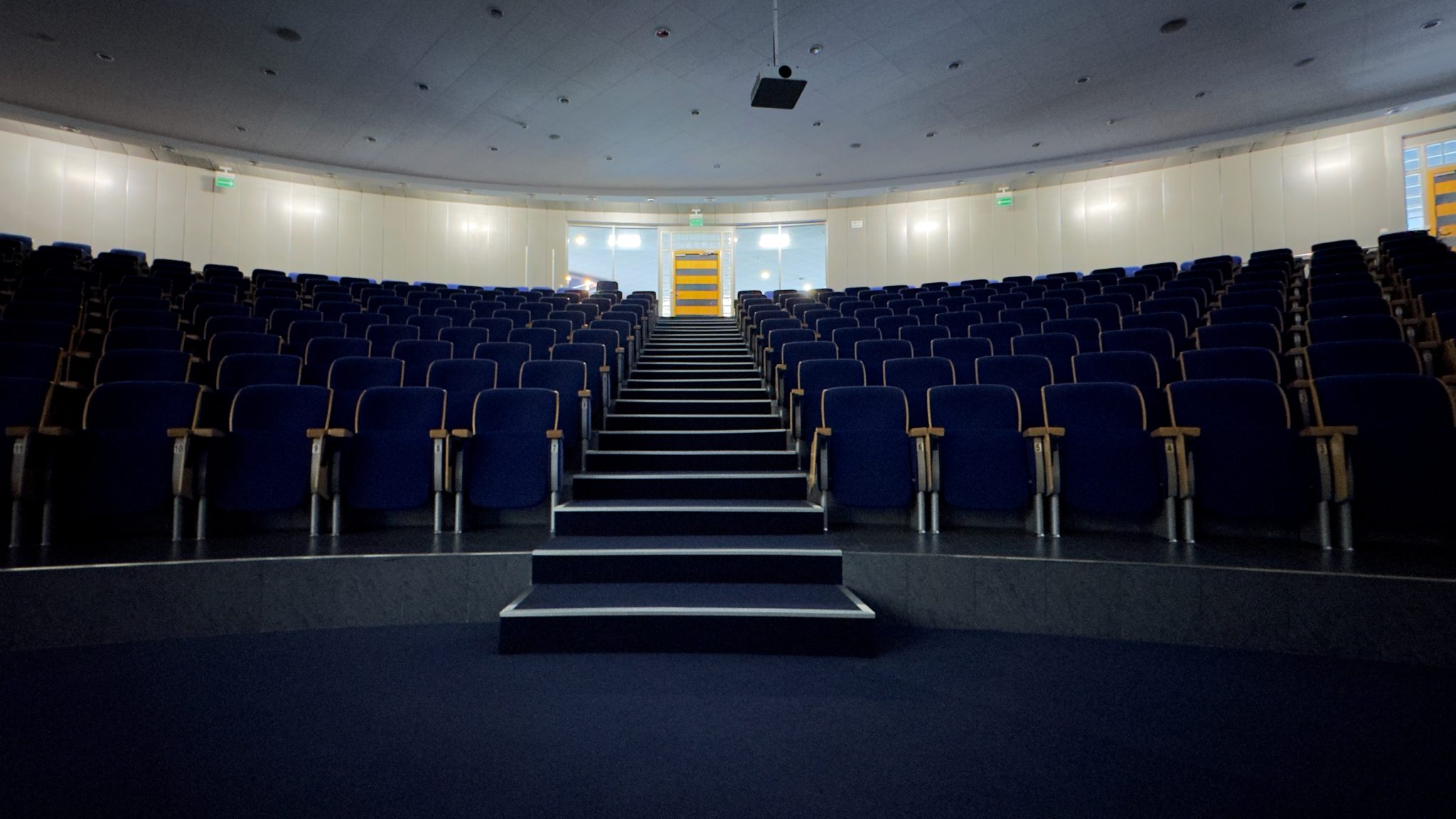 Auditorium. A training room with a circular shape. Blue rows of chairs. A dominant staircase in the center. In the background, the entrance door and a curved wall with lamps. A bright vaulted ceiling. A frog-like perspective.