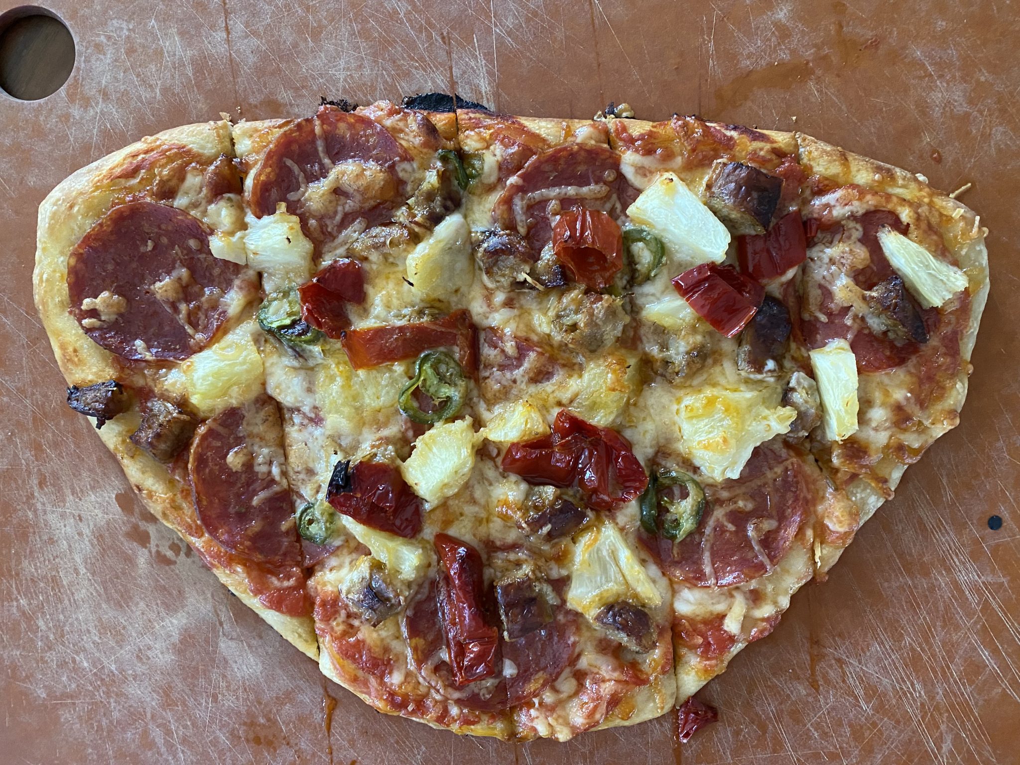 Hand-tossed thin-crust pizza on cutting board, with cheese, pepperoni, sausage, goat horn pepper, pineapple, and jalapeño